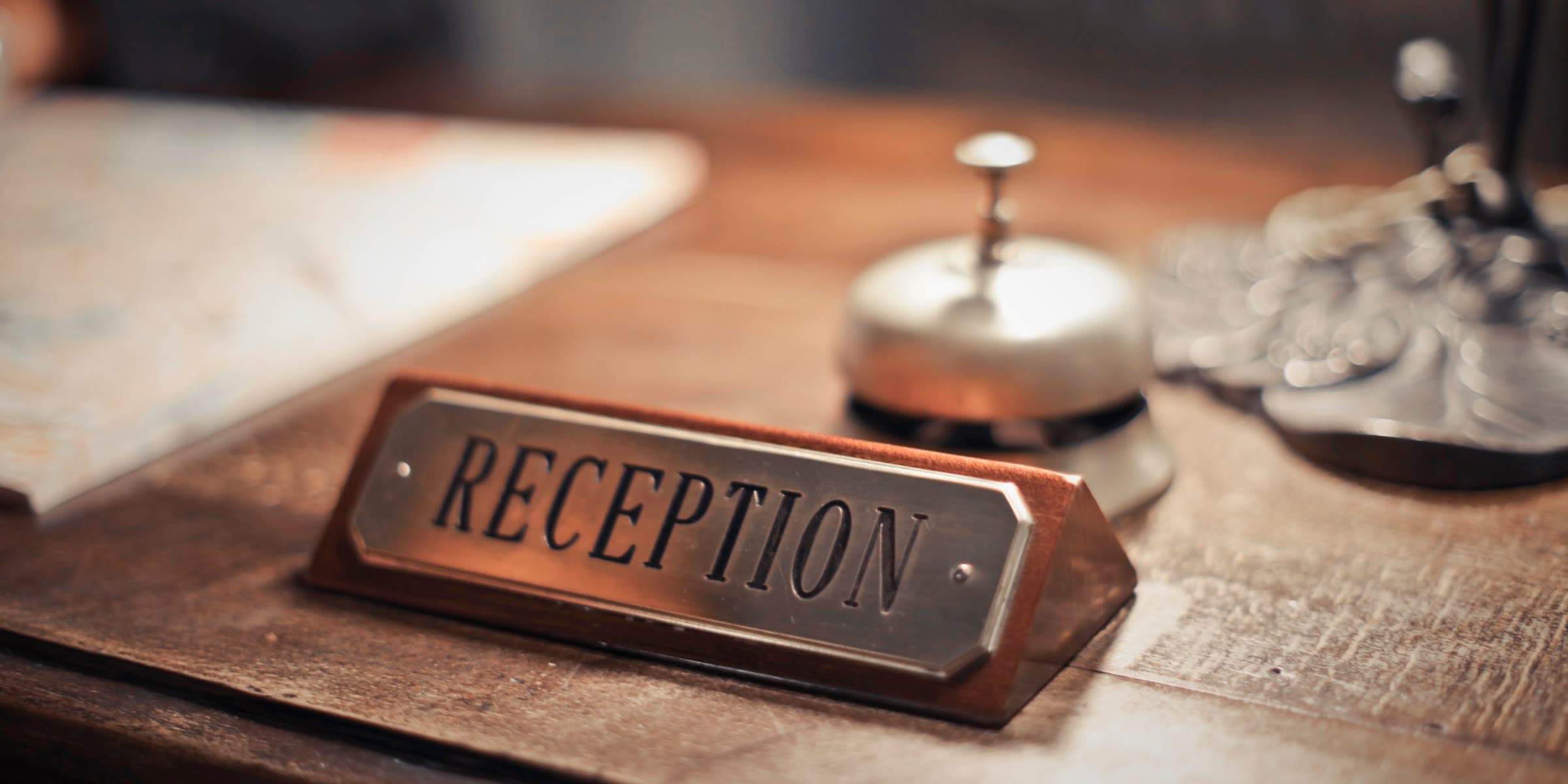 تابلوی برنجی «RECEPTION» و زنگ رومیزی روی پیشخوان چوبی یک هتل، در نور گرم